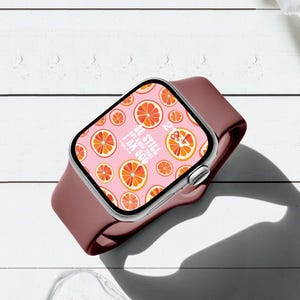 May include: A smartwatch with a dark maroon silicone band and a digital display featuring a repeating pattern of orange citrus slices on a light pink background, with the white text "BE STILL AND KNOW THAT I AM GOD" and a digital clock display.