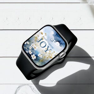 Op de afbeelding: Een zilveren smartwatch met een zwarte band toont een bloemenbehang met het woord "JOY" en de zin "TODAY I CHOOSE". De tijd is 21:57. Het horloge ligt op een wit houten oppervlak.