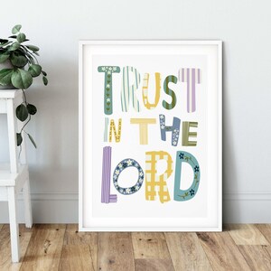 May include: A framed art print featuring the words "TRUST IN THE LORD" in a variety of colourful, patterned fonts. The letters are decorated with stripes, flowers, and plaid patterns. The print is in a white frame, displayed on a wooden floor.