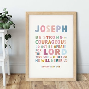 May include: A framed print featuring the name "JOSEPH" in multi-coloured letters. The text below reads "BE STRONG AND COURAGEOUS, DO NOT BE AFRAID FOR THE LORD, YOUR GOD IS WITH YOU; HE WILL NEVER LEAVE YOU" and "DEUTERONOMY 31:6".