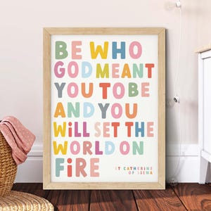 May include: A framed print with the words "BE WHO GOD MEANT YOU TO BE AND YOU WILL SET THE WORLD ON FIRE" in colourful block letters. The print also includes "ST CATHERINE OF SIENA" at the bottom. The frame is light brown.