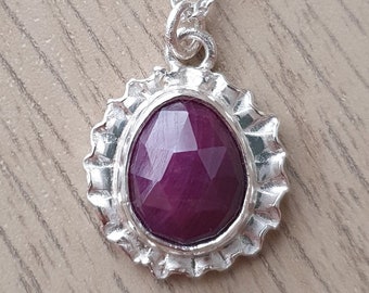 May include: A silver necklace with a pendant featuring a faceted ruby gemstone set in a silver frame. The frame has a scalloped edge.