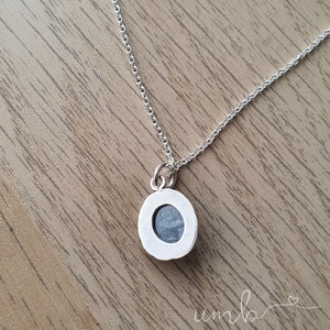 Puede incluir: Un collar de plata con un peque&ntilde;o colgante redondo. El colgante presenta una piedra azul claro engastada en un bisel de plata. La cadena es delicada y el collar se exhibe sobre una superficie de madera.