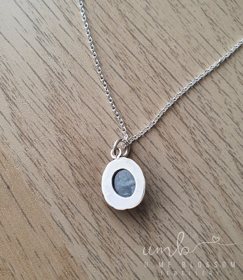 Puede incluir: Un collar de plata con un peque&ntilde;o colgante redondo. El colgante presenta una piedra azul claro engastada en un bisel de plata. La cadena es delicada y el collar se exhibe sobre una superficie de madera.