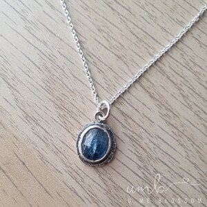 Puede incluir: Un collar de plata con un colgante de piedra preciosa azul. El colgante ovalado est&aacute; engastado en un marco de plata texturizada. La delicada cadena descansa sobre una superficie de madera. Joyer&iacute;a de Ume Blossom Jewellery.