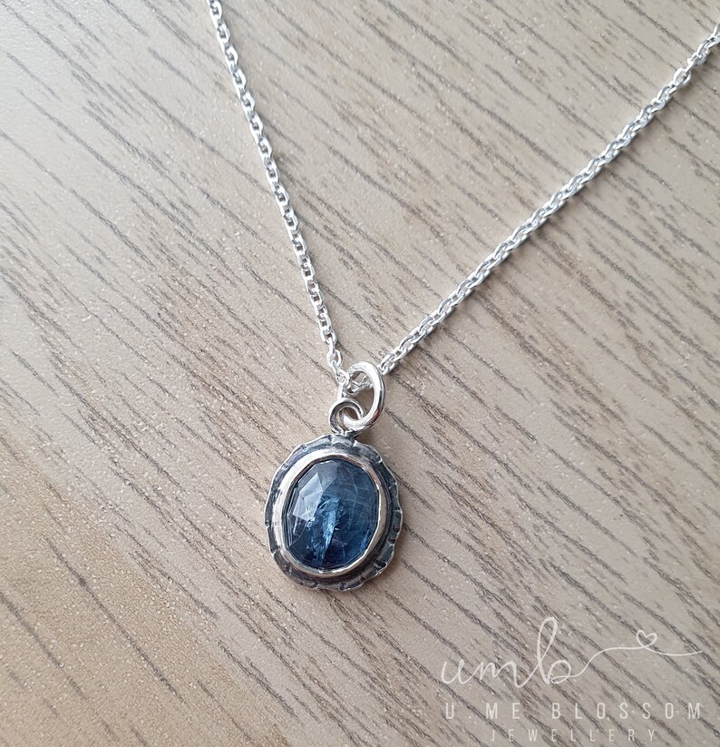 Puede incluir: Un collar de plata con un colgante de piedra preciosa azul. El colgante ovalado est&aacute; engastado en un marco de plata texturizada. La delicada cadena descansa sobre una superficie de madera. Joyer&iacute;a de Ume Blossom Jewellery.