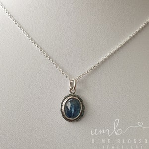 Puede incluir: Un collar de plata con un colgante de piedra preciosa azul. El colgante ovalado est&aacute; engastado en un marco de plata texturizada y cuelga de una delicada cadena. La imagen est&aacute; bien iluminada, mostrando los detalles de la joya. El texto "UMB UME BLOSSOM JEWELLERY" es visible.