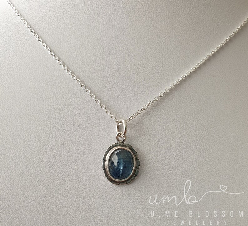 Puede incluir: Un collar de plata con un colgante de piedra preciosa azul. El colgante ovalado est&aacute; engastado en un marco de plata texturizada y cuelga de una delicada cadena. La imagen est&aacute; bien iluminada, mostrando los detalles de la joya. El texto "UMB UME BLOSSOM JEWELLERY" es visible.