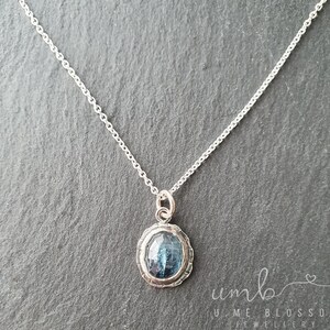 Puede incluir: Un collar de plata con un colgante de piedra preciosa azul. El colgante ovalado tiene un borde plateado texturizado y una piedra azul facetada. La cadena es delicada y plateada. El collar se muestra sobre una superficie gris oscuro.