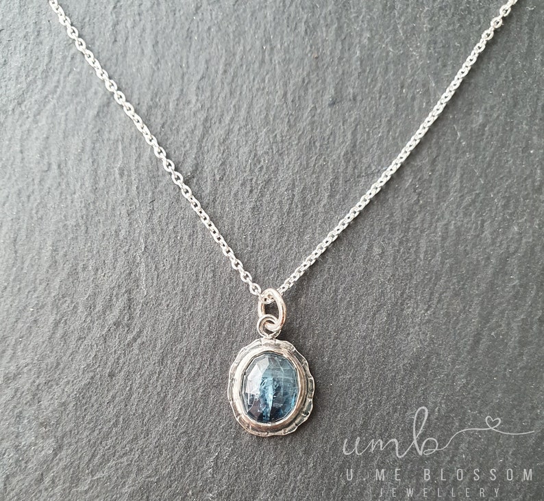 Puede incluir: Un collar de plata con un colgante de piedra preciosa azul. El colgante ovalado tiene un borde plateado texturizado y una piedra azul facetada. La cadena es delicada y plateada. El collar se muestra sobre una superficie gris oscuro.
