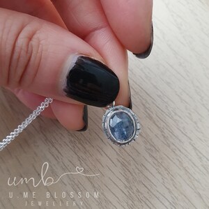 Puede incluir: Un collar de plata con un colgante de piedra azul. El colgante ovalado est&aacute; engastado en un marco de plata texturizada. El collar lo sostiene una persona con esmalte de u&ntilde;as negro. La marca "U. ME BLOSSOM JEWELLERY" es visible.
