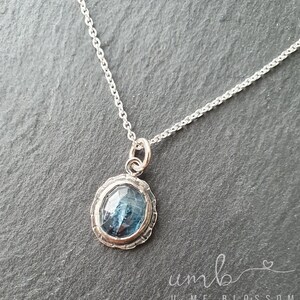 Puede incluir: Un collar de plata con un colgante de piedra preciosa azul. El colgante tiene forma ovalada con un borde plateado texturizado y una piedra azul facetada. La cadena es delicada y plateada.