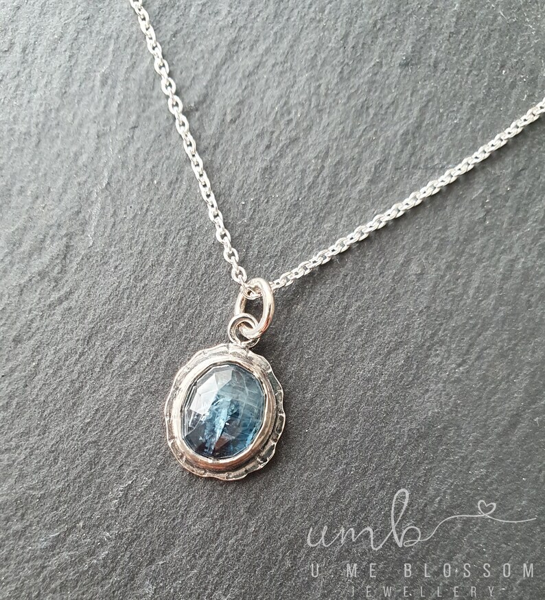 Puede incluir: Un collar de plata con un colgante de piedra preciosa azul. El colgante tiene forma ovalada con un borde plateado texturizado y una piedra azul facetada. La cadena es delicada y plateada.