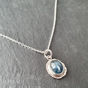 Puede incluir: Un collar de plata con un colgante de piedra preciosa azul. El colgante ovalado tiene una piedra azul facetada engastada en un bisel de plata texturizada. La cadena es de estilo eslabones delicados. El collar se muestra sobre una superficie gris oscuro.