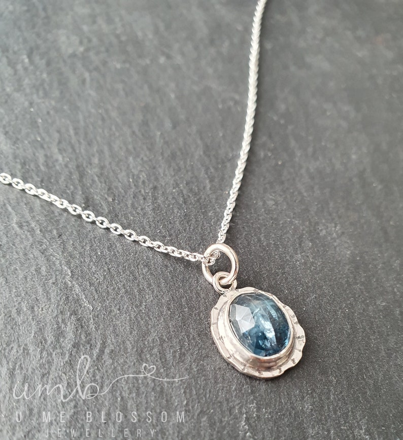 Puede incluir: Un collar de plata con un colgante de piedra preciosa azul. El colgante ovalado tiene una piedra azul facetada engastada en un bisel de plata texturizada. La cadena es de estilo eslabones delicados. El collar se muestra sobre una superficie gris oscuro.