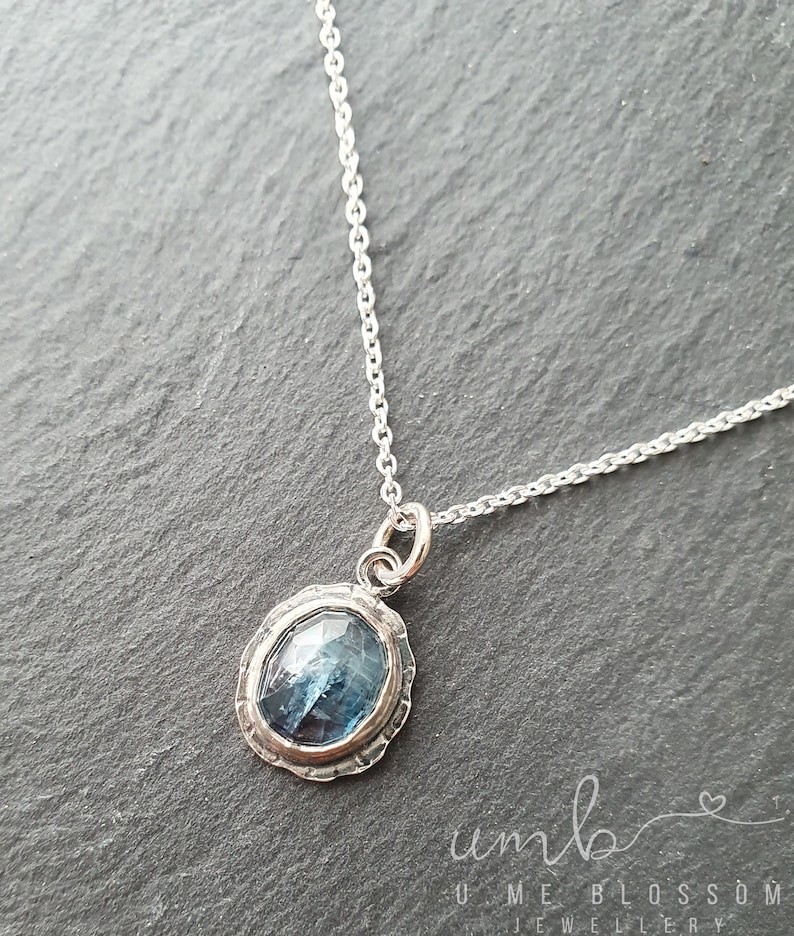 Puede incluir: Un collar de plata con un colgante de piedra azul. El colgante ovalado tiene un borde plateado texturizado y una piedra azul facetada. La delicada cadena sostiene el colgante desde un peque&ntilde;o lazo. El fondo es una superficie gris oscuro.