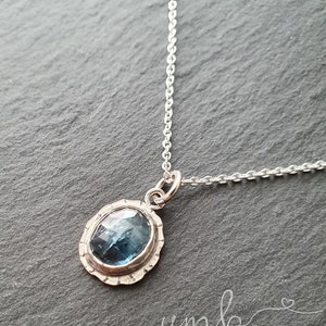 Puede incluir: Un collar con colgante de plata con una piedra preciosa azul ovalada. La piedra preciosa est&aacute; engastada en un bisel festoneado texturizado. La delicada cadena de plata es visible. La joya se exhibe sobre una superficie gris oscuro.