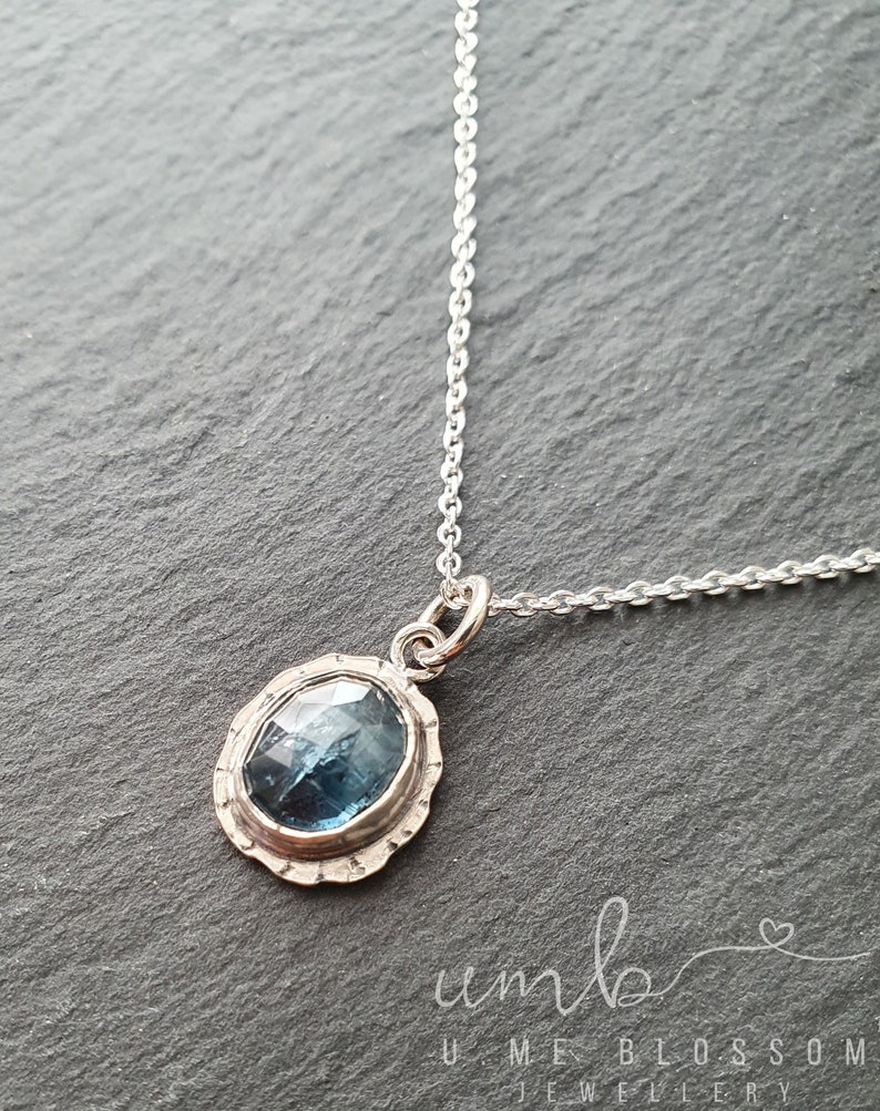 Puede incluir: Un collar con colgante de plata con una piedra preciosa azul ovalada. La piedra preciosa est&aacute; engastada en un bisel festoneado texturizado. La delicada cadena de plata es visible. La joya se exhibe sobre una superficie gris oscuro.