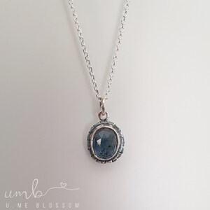 Puede incluir: Un collar de plata con un colgante ovalado azul. El colgante tiene una piedra azul oscura texturizada engastada en un marco de plata decorativo. La delicada cadena cuelga sobre un fondo blanco. El collar es una joya.