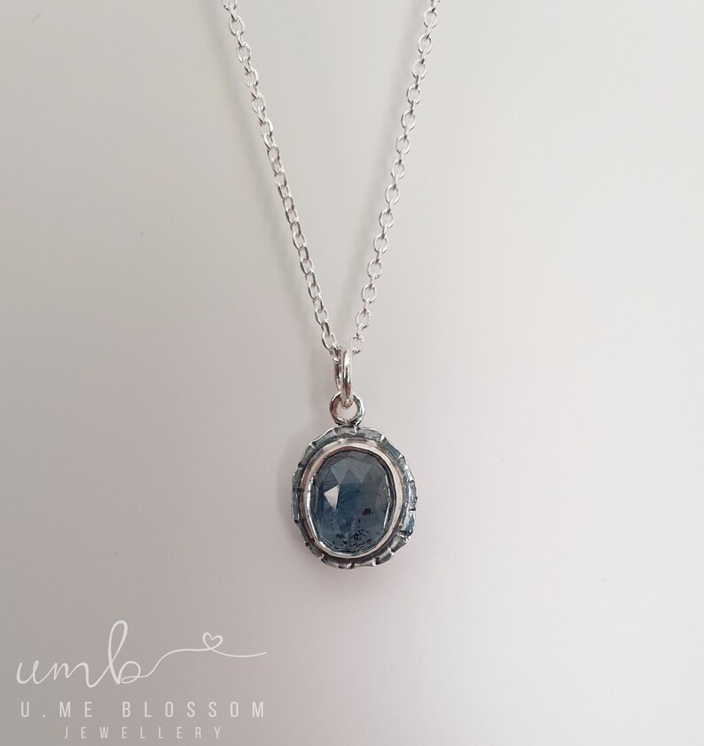 Puede incluir: Un collar de plata con un colgante ovalado azul. El colgante tiene una piedra azul oscura texturizada engastada en un marco de plata decorativo. La delicada cadena cuelga sobre un fondo blanco. El collar es una joya.
