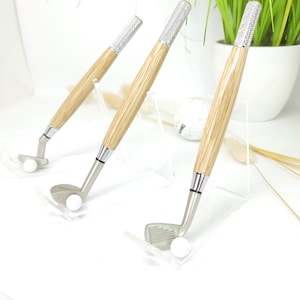May include: Three golf club-shaped pens with wooden barrels and silver club heads. The pen tops are encrusted with rhinestones. Each pen has a white golf ball in front of the club head. The pens are displayed on a white surface.