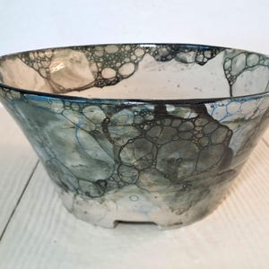 Può includere: Una ciotola in ceramica caratterizzata da un motivo a smalto a bolle in tonalità verde scuro, blu e nero su base bianca. La superficie esterna presenta una complessa rete di forme circolari interconnesse, creando un effetto visivo strutturato.