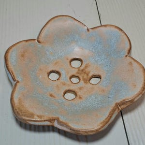 May include: A ceramic soap dish in the shape of a flower. The dish has a light blue and tan color scheme with five drainage holes. The edges are a darker brown. The dish is designed to hold a bar of soap.