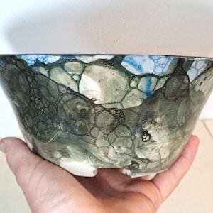 May include: A ceramic bowl featuring a dark green and blue bubble-like pattern across its exterior surface. The glaze displays a complex network of interconnected circular shapes and dark outlines, with a smooth, glossy finish and a white base.