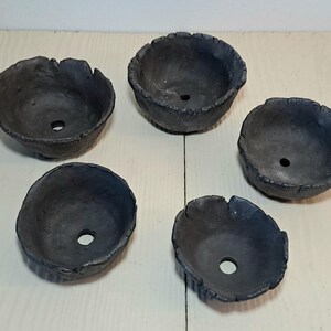 Peut inclure: Cinq bols en céramique gris foncé aux bords irréguliers et texturés. Chaque bol a un trou de drainage circulaire au fond. Les bols sont disposés sur une surface en bois de couleur claire.