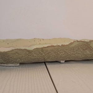 May include: An elongated, rectangular ceramic tray with a light beige, off-white finish. The tray features a textured, embossed pattern along its exterior sides and a wavy, uneven rim. It is supported by small, low-profile feet at the base.