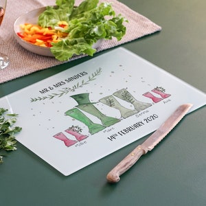 Puede incluir: Una tabla de cortar de vidrio blanco con un diseño personalizado que presenta una familia de botas de lluvia verdes y rosas con nombres y la fecha "14 de febrero de 2020". El texto "Mr & Mrs Sanders" está en la parte superior de la tabla.