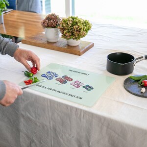 May include: A glass cutting board with a light blue background and a white border. The board has a design of flip-flops with different patterns and the text "The Grilley Family" and "Noodstyle Surfing and Sun Since 2016". A person is using a knife to cut red and green peppers on the board.