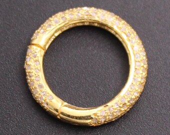 May include: A gold-colored, circular clasp with a hinged opening. The clasp is encrusted with small, clear gemstones, creating a sparkling effect. The background is a solid, dark gray color.