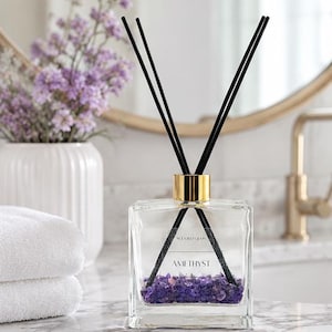 May include: A clear square glass reed diffuser bottle with a gold-colored collar and black reeds. The bottle contains small purple amethyst stones at the bottom and a label that reads SCENTED GEMS AMETHYST in black text on the front surface.