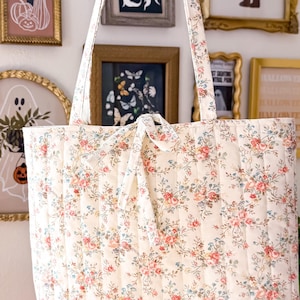 May include: A quilted tote bag featuring a cream-colored fabric with a delicate floral pattern in shades of pink, blue, and green. The bag has vertical stitching, two shoulder straps, and a small fabric tie closure at the top center.