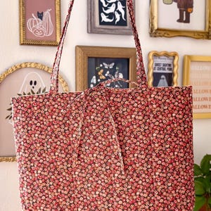 May include: A quilted tote bag featuring a dense, repeating pattern of small red, pink, and white flowers on a dark background. The bag has two long shoulder straps and a tie closure at the top center, constructed with vertical stitching lines.