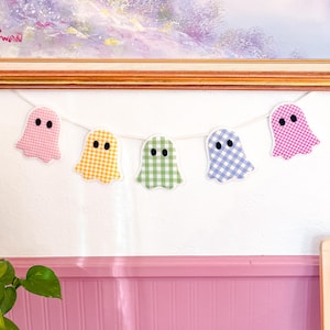 May include: A decorative garland featuring five ghost-shaped cutouts suspended on a white string. Each ghost is made of fabric with a gingham check pattern in pastel pink, yellow, light green, light blue, and magenta, each with two black eyes.