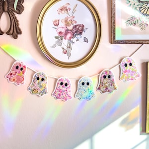 May include: A decorative garland featuring six ghost-shaped ornaments strung along a white cord. Each ghost is crafted from fabric with various floral patterns in shades of pink, yellow, blue, and green, and features two black felt oval eyes.