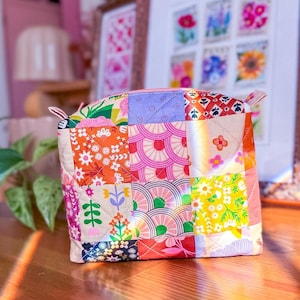 May include: A colorful patchwork pouch with a pink zipper and small fabric loops. The fabric is a mix of floral and geometric patterns in shades of orange, pink, yellow, and green. The pouch is sitting on a wooden surface.