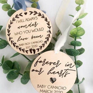 May include: Two wooden ornaments with engraved text. The top ornament is light brown and features the text "I will always wonder who you would have been" with a small heart and floral design. The bottom ornament is a darker brown and features the text "forever in our hearts" with a small heart and the text "Baby Camacho March 2020".