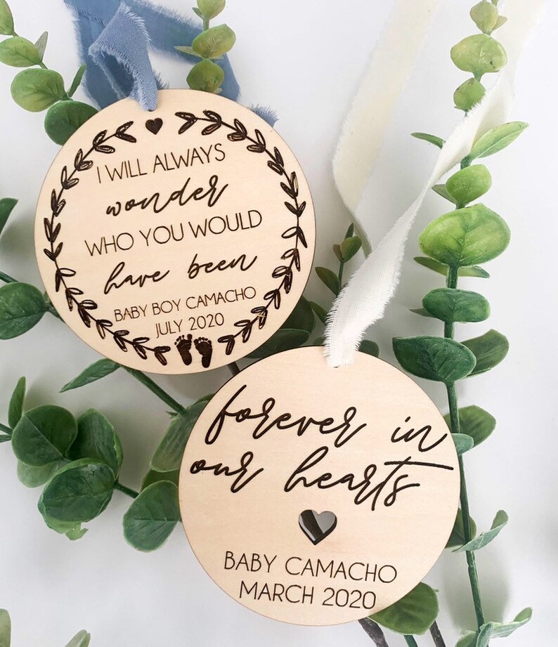 May include: Two wooden ornaments with engraved text. The top ornament is light brown and features the text "I will always wonder who you would have been" with a small heart and floral design. The bottom ornament is a darker brown and features the text "forever in our hearts" with a small heart and the text "Baby Camacho March 2020".