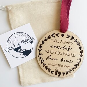May include: A wooden ornament with the text "I will always wonder who you would have been" and the name "Taylor Lee Logan" and the year "2019". The ornament is decorated with a heart and a wreath of leaves.