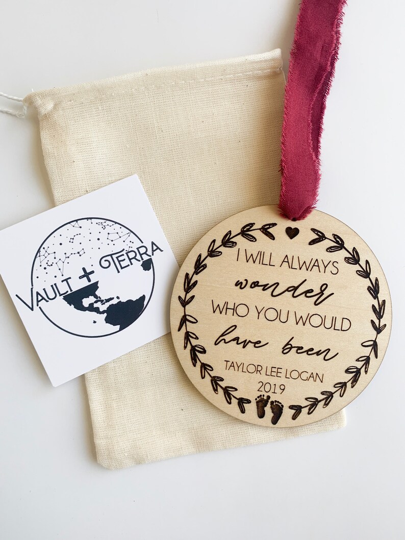 May include: A wooden ornament with the text "I will always wonder who you would have been" and the name "Taylor Lee Logan" and the year "2019". The ornament is decorated with a heart and a wreath of leaves.