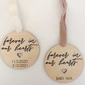 May include: Two wooden ornaments with the text "forever in our hearts" and a heart cutout. One ornament has the dates "11.5.2020" and "3.10.2021" printed on it. The other ornament has the text "BABY AVA" printed on it.