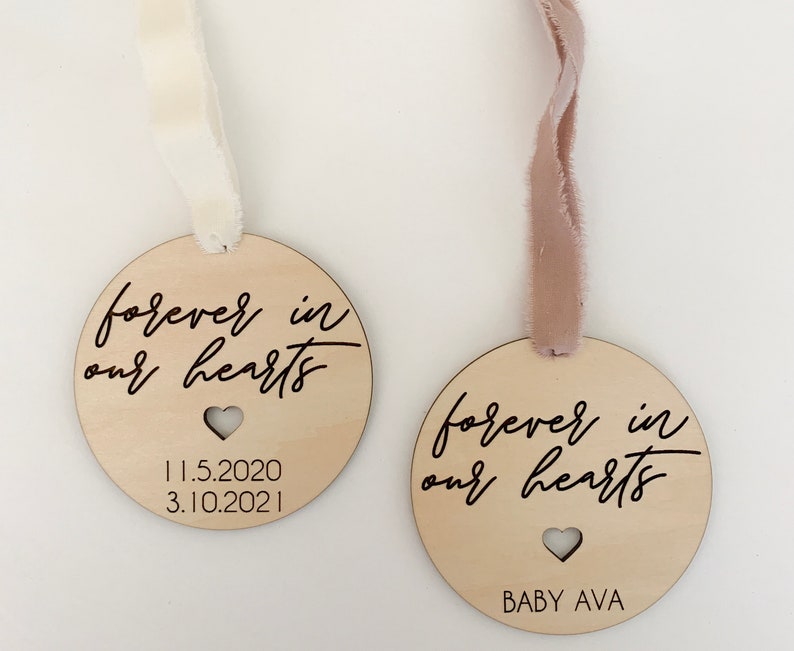 May include: Two wooden ornaments with the text "forever in our hearts" and a heart cutout. One ornament has the dates "11.5.2020" and "3.10.2021" printed on it. The other ornament has the text "BABY AVA" printed on it.