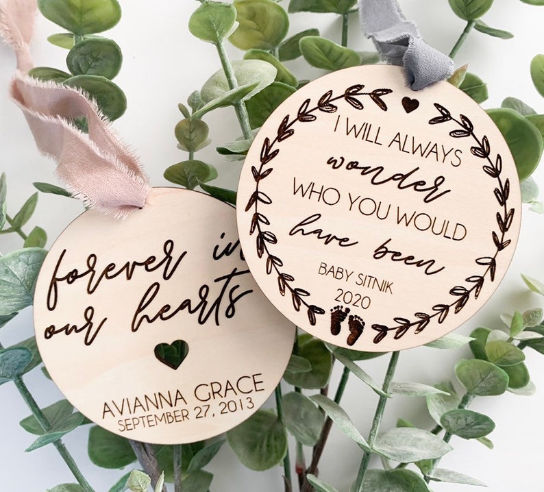 May include: Two wooden ornaments with engraved text. The first ornament reads "forever in our hearts" with the name "AVIANNA GRACE" and the date "SEPTEMBER 27, 2013". The second ornament reads "I WILL ALWAYS wonder WHO YOU WOULD have been" with the name "BABY SITNIK" and the date "2020".