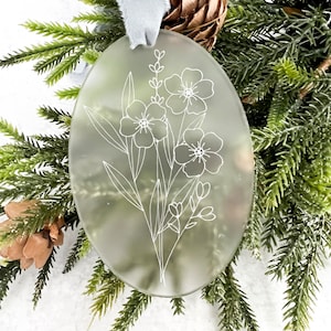 Puede incluir: Un adorno ovalado de vidrio esmerilado con un diseño floral blanco. El adorno está colgado de una cinta azul.