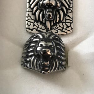 May include: A silver-toned metal ring featuring a sculpted lion head with an open mouth and a matching rectangular pendant with a similar lion head design set against a textured, irregular background pattern of raised silver-colored shapes.