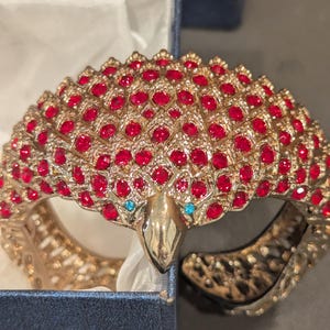 May include: Gold-toned metal bracelet with a peacock design. The bracelet is encrusted with numerous red rhinestones and features a peacock head with turquoise-colored eyes. The bracelet is presented in a blue box.