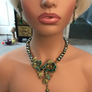 May include: A necklace and earring set featuring a gold-tone metal snake motif adorned with green crystals and beads. The necklace includes a central snake pendant with a dangling teardrop-shaped green crystal, while the earrings are matching drops.